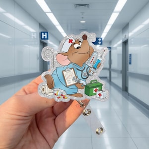 May include: A glittery badge reel featuring a cartoon mouse dressed as a nurse, holding a clipboard, syringe, and thermometer. The mouse wears a nurse's cap with a red cross. A green first aid kit and pills are also visible.