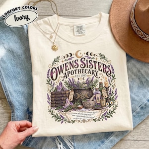 May include: An ivory t-shirt featuring a graphic print of a cauldron, stacked books, lavender sprigs, and a candle. The text reads "Owens Sisters Apothecary Est. 1995" along with a quote about rosemary and lavender. The design includes moon phases.