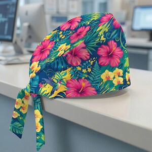 May include: A scrub cap featuring a vibrant tropical pattern on a dark blue background. The design includes bright pink hibiscus flowers, yellow orchids, green fern leaves, and a small hummingbird. The cap has two fabric ties hanging from the back.