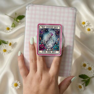 May include: A rectangular phone grip featuring a graphic illustration of a skeleton hand holding a smoking sage bundle, surrounded by colorful flowers and stars. The text "NOT ENOUGH SAGE FOR THIS SHIT" is printed in black on a cream background.