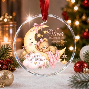 Puede incluir: Un adorno de cristal circular con una cinta roja que muestra un osito de peluche durmiendo sobre una luna creciente amarilla, flores rosas y una nube con el texto "HAPPY FIRST BIRTHDAY". Incluye el nombre "Oliver", "17th AUGUST 2025" y "Love from Auntie Emma".