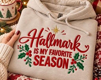 May include: A light gray hooded sweatshirt featuring red embroidered text that reads Hallmark IS MY FAVORITE SEASON, accented with a gold star and green holly leaves with red berries on the front center.