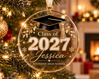 May include: A clear circular ornament featuring a black graduation cap with a gold tassel, the text Class of 2027, the name Jessica, and Riverside High School. The design includes gold leaf accents and white snowflakes, suspended by a white ribbon.