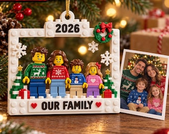 Puede incluir: Un adorno de marco hecho con bloques de construcción blancos con el texto 2026 y OUR FAMILY. Cuatro minifiguras con suéteres navideños están dentro del marco, flanqueadas por pequeños árboles de Navidad de bloques. Una foto familiar está al lado.