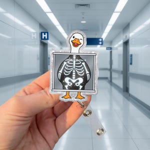 Pode incluir: Um porta-crachá com um pato de desenho animado piscando, com um raio-x de seu esqueleto. O porta-crachá tem uma alça de plástico transparente e uma borda prateada com glitter. O pato tem pés laranja e um corpo branco.