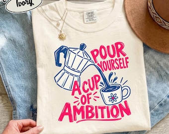 May include: An ivory t-shirt featuring a graphic of a blue moka pot pouring coffee into a cup. The text reads "POUR YOURSELF A CUP OF AMBITION" in pink and blue lettering. The shirt has a Comfort Colors tag and is paired with blue denim.