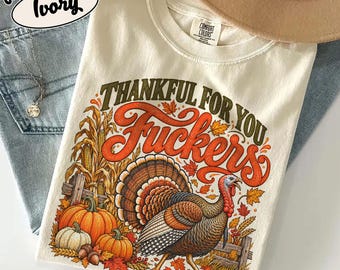 May include: An ivory t-shirt featuring a graphic of a turkey, pumpkins, and corn stalks. The text reads Thankful For You Fuckers in orange and dark green script. The shirt has a Comfort Colors label at the neck and is displayed on blue denim.