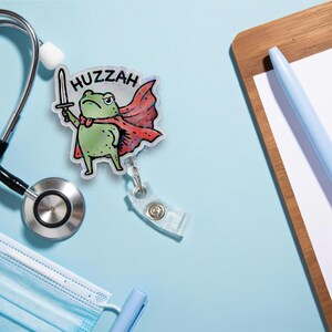 May include: A badge reel featuring a cartoon frog in a red cape holding a sword, with the word "Huzzah" above it. The badge reel is on a light blue surface with a stethoscope, face mask, pen, and notepad.