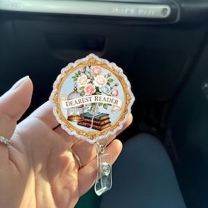May include: A round badge reel with a floral design and the words "Dearest Reader" on a banner. The badge reel features a bouquet of pink, white, and blue roses, a birdcage, and a stack of books. The reel has a clear plastic clip.