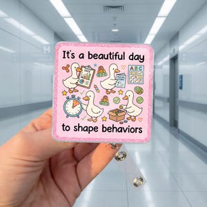 May include: A square badge reel with a pink glitter border featuring four white cartoon ducks and the text It's a beautiful day to shape behaviors. The design includes illustrations of a data chart, a social story book, an ABC chart, a clock, and a box.