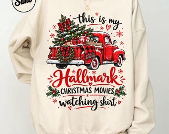 May include: A sand-colored crewneck sweatshirt featuring a graphic of a red pickup truck carrying a Christmas tree and gifts. The text reads, "this is my Hallmark CHRISTMAS MOVIES watching shirt," surrounded by red snowflakes and hearts.