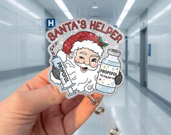 以下が含まれることがあります： PROPOFOLと書かれた注射器とPROPOFOL 10 mg per mlと書かれたバイアルを持つ、ウィンクしたサンタクロースのイラストが描かれたグリッターバッジリール。キャラクターの上には赤い文字でSANTA'S HELPERとアーチ状に書かれています。