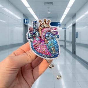 May include: A badge reel featuring a colorful anatomical heart design. The heart is labeled with anatomical terms like "aorta" and "ventricle." The design includes a leopard print pattern and a blue bow. The reel has a clear plastic strap.