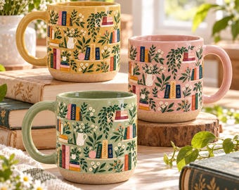 May include: Three ceramic mugs in yellow, pink, and sage green, each featuring a wrap-around illustration of bookshelves filled with colorful books, potted plants, and small white flowers. Each mug has a speckled, unglazed base and a handle.