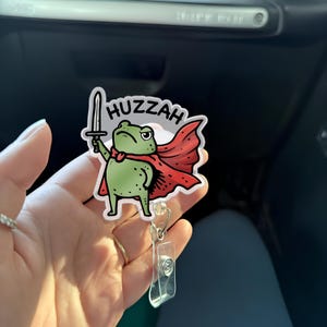 May include: A green frog character with a red cape and a sword, the word "Huzzah" is above the frog. The frog is a badge reel with a clear plastic clip and retractable cord.