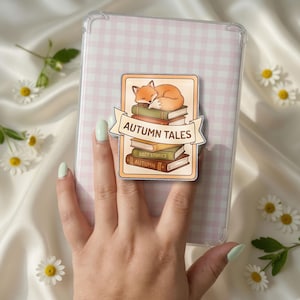 May include: A phone grip featuring a rectangular illustration of a sleeping orange fox atop a stack of books. A cream-colored banner across the center displays the text "AUTUMN TALES" in brown. The books are labeled "COZY STORIES" and "AUTUMN".