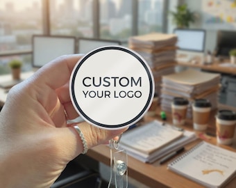 May include: A round badge reel featuring a white circular face with a black border and the black text CUSTOM YOUR LOGO printed in the center. The badge reel has a clear plastic strap with a metal snap button attachment for holding identification cards.