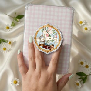 May include: A clear phone grip featuring a circular gold-trimmed frame with a scalloped edge. Inside the frame is an illustration of a bouquet of pink and white roses, a birdcage, and a stack of books with the text DEAREST READER on a banner.