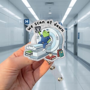 May include: A glittery acrylic badge reel featuring a cartoon frog in blue scrubs sitting on a CT scanner, holding an X-ray film. The text "we scan at dawn" is printed above the frog. Several books and a coffee mug are placed near the scanner base.