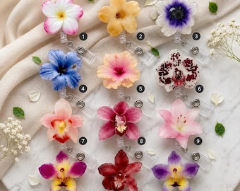 Puede incluir: Una colección de doce portatarjetas, cada uno con un diseño floral único. Las flores incluyen varias orquídeas, hibiscos y flores tipo anémona en tonos rosa, azul, amarillo, morado, blanco y rojo oscuro, todas unidas a clips transparentes.
