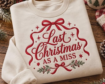 May include: A cream-colored crewneck sweatshirt featuring red embroidered text that reads Last Christmas AS A MISS. The design includes a red ribbon bow at the top and bottom, small red stars, and green holly leaves with red berries.