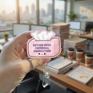 May include: A pink badge reel featuring a tissue box design with the text "Crying Over Fictional Characters." The tissue box has a cute face and star accents. The reel has a clear plastic clip.