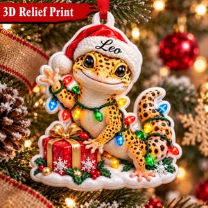 Op de afbeelding: Een 3D-reliëf kerstornament met een geel-zwart gevlekte luipaardgekko die een rood-witte kerstmuts draagt. De gekko is omwikkeld met kleurrijke kerstverlichting naast een rood cadeau met een gouden strik.