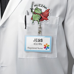 May include: A white lab coat with a pocket holding a pen and a name badge. A cartoon frog with a sword and cape is attached to the coat. The name badge reads "JESS ICU RN Registered Nurse". The frog has the word "Huzzah" above it.
