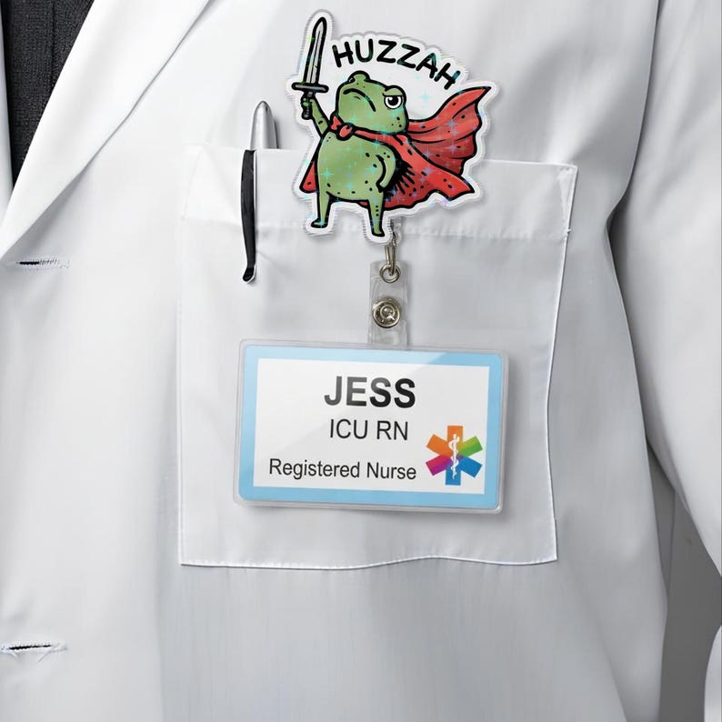 May include: A white lab coat with a pocket holding a pen and a name badge. A cartoon frog with a sword and cape is attached to the coat. The name badge reads "JESS ICU RN Registered Nurse". The frog has the word "Huzzah" above it.