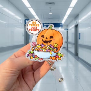 May include: A glittery acrylic badge reel featuring a smiling orange jack-o'-lantern holding a white bowl filled with colorful assorted candies. A speech bubble above the pumpkin displays the orange text NO TRICKS JUST TREATS in capital letters.