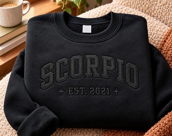 May include: A black crewneck sweatshirt featuring the word SCORPIO in a large, arched, dark-toned embroidered font across the chest, with the text EST. 2021 centered below, flanked by two small four-pointed star symbols.