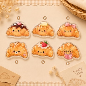 May include: Six acrylic charms shaped like golden brown croissants with cute faces. Designs include a chocolate-topped croissant with a heart, a buttered one, a bow-adorned one, a bear-shaped one, a strawberry-topped one, and one reading a book.