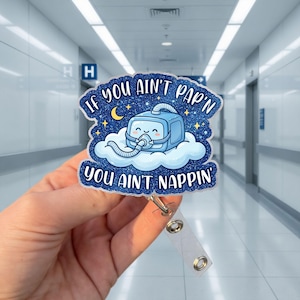 Peut inclure: Un porte-badge bleu pailleté représentant une machine CPAP de dessin animé avec un visage souriant reposant sur un nuage blanc. Le design inclut un croissant de lune jaune et des étoiles sur fond bleu foncé avec le texte IF YOU AIN'T PAP'N YOU AIN'T NAPPIN'.
