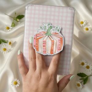 Op de afbeelding: Een ronde telefoonhouder met een grafische illustratie van een pompoen gemaakt van open boeken. De pompoen is oranje en roze met een groene steel en rankdetails. De houder is bevestigd aan een tablethoes met een roze en wit vichy-ruitpatroon.