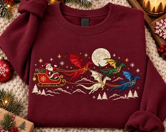May include: A maroon crewneck sweatshirt featuring a graphic print of Santa Claus in a sleigh pulled by four dragons in red, green, white, and blue colors. The design includes a full moon, stars, and snowy pine trees against a dark background.