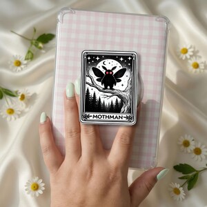 May include: A rectangular phone grip featuring a black and white illustration of the Mothman with glowing red eyes, perched on a tree branch under a moon and stars, with the text MOTHMAN printed at the bottom, attached to a pink gingham case.