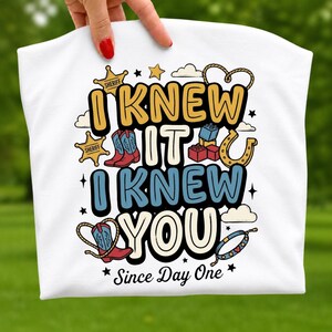 May include: A white t-shirt featuring a graphic design with the text I KNEW IT I KNEW YOU Since Day One. The illustration includes yellow sheriff stars, red and blue cowboy boots, building blocks, a horseshoe, a lasso, and a beaded bracelet.