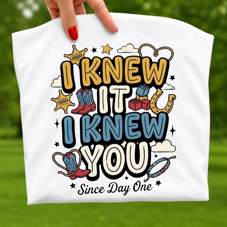 May include: A white t-shirt featuring a graphic design with the text I KNEW IT I KNEW YOU Since Day One. The illustration includes yellow sheriff stars, red and blue cowboy boots, building blocks, a horseshoe, a lasso, and a beaded bracelet.