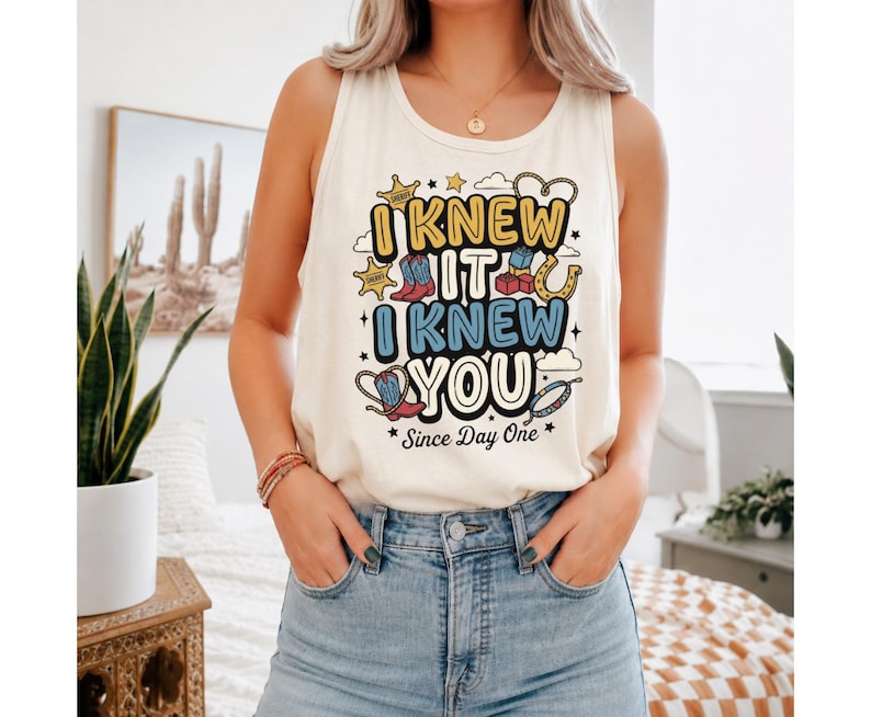 May include: A cream-colored sleeveless tank top featuring a graphic print with the text "I KNEW IT I KNEW YOU Since Day One" in yellow and blue. The design includes illustrations of cowboy boots, sheriff stars, and a horseshoe.