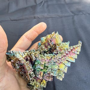 May include: A colorful bismuth crystal with a geometric, layered structure. The crystal displays a vibrant array of colors including blues, yellows, greens, and purples. The mineral is held in a hand.