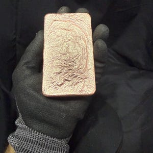 May include: A rectangular copper bar with a textured surface is held by a gloved hand. The bar has a reddish-brown color and a unique, organic pattern across its surface. The background is dark, emphasizing the bar's details.