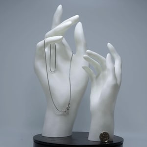 Pode incluir: Um colar de prata exibido em um suporte de joias branco em forma de mão. O colar é delicado e pende com graça. O suporte de joias está sobre uma base preta, com uma moeda por perto. O fundo é cinza suave.