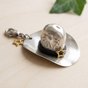 May include: A silver-colored cowboy hat keychain with a decorative star charm. The hat features a detailed design and a black band. The keychain includes a clasp for easy attachment.