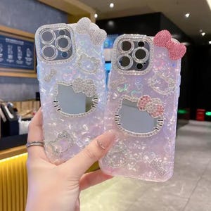 Puede incluir: Dos fundas de tel&eacute;fono iridiscentes con dise&ntilde;os de Hello Kitty. Cada funda presenta un lazo plateado, una cara de Hello Kitty y adornos brillantes. Las fundas est&aacute;n en la mano de una persona.