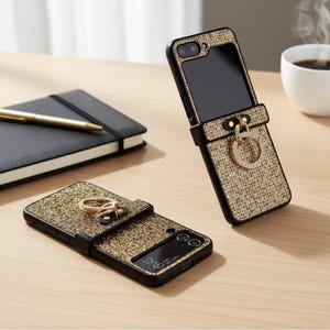 May include: Two black and gold phone cases with a textured, glittery design. One case is open, revealing the phone screen, and features a ring holder. The other case is closed. A black notebook, pen, and coffee cup are in the background.