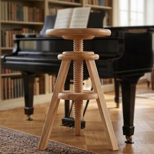 May include: A light-colored wooden piano stool with a round seat and a threaded height adjustment mechanism. The stool has three angled legs and is positioned in front of a black grand piano. A bookshelf filled with books is in the background.
