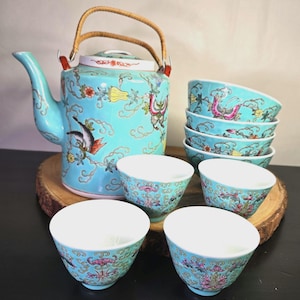 May include: A turquoise ceramic tea set featuring a teapot with a woven handle and six matching small cups. The exterior of each piece is decorated with colorful floral patterns, butterflies, and swirling line motifs against a bright blue background.
