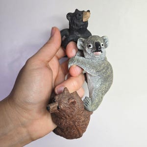 May include: Three small animal figurines including a black bear with a tan snout, a grey koala with a black nose, and a brown bear. Each figurine is sculpted with textured fur details and realistic features, designed to hang or grip onto surfaces.