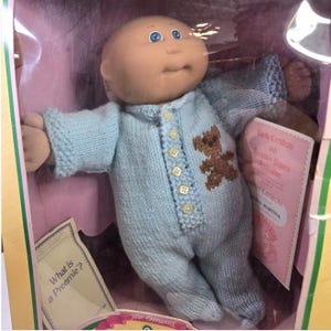 Puede incluir: Muñeca prematura Cabbage Patch Kids en una caja. La muñeca tiene la cabeza calva, ojos azules y lleva un mono de punto azul claro con una aplicación de oso de peluche marrón. La caja incluye un certificado de nacimiento y otros papeles.