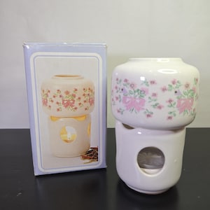 May include: A white ceramic oil warmer featuring a floral pattern with pink bows and green leaves. The warmer consists of a top bowl and a base with a circular opening for a tea light candle. It is displayed next to its original light blue cardboard box.
